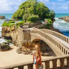 Mujer con mochila admira un puente que cruza sobre el mar, disfrutando de la tranquilidad de la **temporada baja** en la costa rocosa.