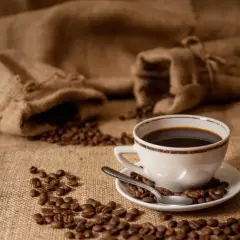 Aromatic coffee beans spill from burlap sacks, surrounding a white cup of dark brew. Zastanawiasz się, ile kofeiny ma kawa parzona?