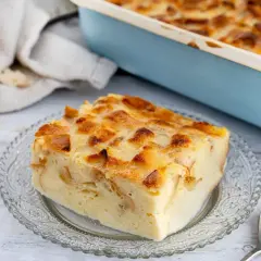 Ein Stück goldbrauner Brotpudding auf einem Glas-Teller, daneben ein Laib Brot und eine Auflaufform.
