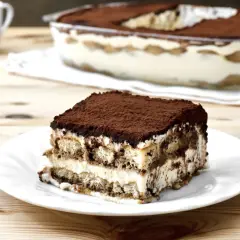 Una fetta di tiramisù su un piatto bianco, con la teglia sullo sfondo. La crema tiramisù perfetta, soffice e stratificata, è cosparsa di cacao.