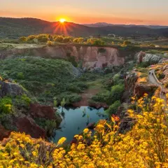 Atardecer dorado sobre un cañón con un estanque, ideal para senderismo Sevilla. Flores amarillas bordean el camino.
