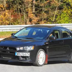 Czarny Mitsubishi Lancer 9 z białymi felgami i czerwonymi chlapacami na tle jesiennego lasu.