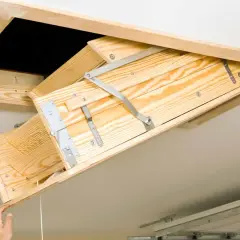 A wooden attic ladder is being installed, with a hand pulling down the first section.