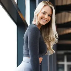 Uśmiechnięta blondynka w szarym sportowym komplecie, z frotką Jordan na nadgarstku, pozuje przy oknie.