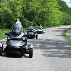 Trzy pojazdy typu Can-Am Spyder jadące po krętej drodze wśród zieleni. Ich nowoczesny design i dynamiczna jazda tworzą wrażenie efektu shimmy.