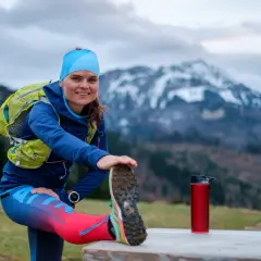 Uśmiechnięta biegaczka przygotowuje się do treningu ultramaraton 50 km, rozciągając nogę na tle gór.
