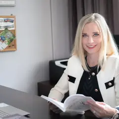 Uśmiechnięta blondynka w białej marynarce siedzi przy biurku i czyta książkę.