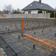 Zbrojenie i rury kanalizacyjne na poziomie zero budynku. W tle dom i ogrodzenie.