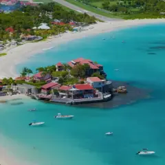 Vue aérienne de Saint-Barthélemy, avec ses plages de sable blanc, ses eaux turquoise et ses villas aux toits rouges. Un voilier navigue paisiblement.