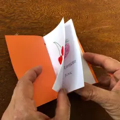 Mani che sfogliano un piccolo libro fatto a mano, con disegni di fragole e ciliegie. Un'idea per imparare come fare un libro a mano.