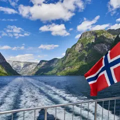 Norweska bandera flaga powiewa na tle malowniczego fiordu. Statek pruje wodę, zostawiając za sobą pianę.