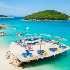 Idealne miejsce, gdzie na weekend nad morze: drewniany pomost z leżakami i parasolami, turkusowa woda, piaszczysta plaża i zielona wyspa w tle.