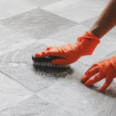 Ręce w pomarańczowych rękawiczkach szorują szczotką podłogę z płytek, pokazując, jak usunąć kamień z płytek łazienkowych.