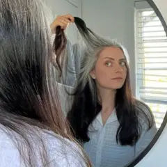 Mujer se mira al espejo, su cabello es una mezcla de negro y canas amarillas, un estilo natural y único.
