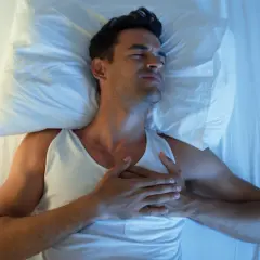 Mann liegt im Bett, hält sich die Brust. Er leidet unter Herzrasen nachts, die Augen sind geschlossen, das Gesicht verzogen.