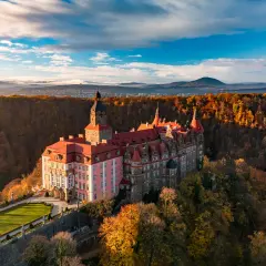 Malowniczy hotel zamek otoczony jesiennym lasem, z wieżą i czerwonymi dachami, na tle gór i błękitnego nieba.