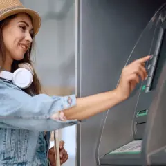 Uśmiechnięta kobieta w kapeluszu i dżinsowej kurtce dokonuje wypłaty z bankomatu za granicą.
