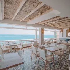 Comedor de la casa Pepe San José con vistas al mar. Mesas puestas, sillas blancas y techo de caña.