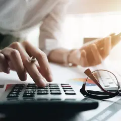 Dłoń naciska klawisze kalkulatora, sprawdzając, czy można dobrać kredyt gotówkowy. Obok leżą okulary i telefon.