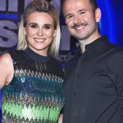 Uśmiechnięci Sławomir i Kajra pozują do zdjęcia na tle niebieskiej kurtyny. Kajra ma na sobie błyszczącą sukienkę, a Sławomir czarną koszulę.