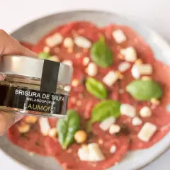 Mano sostiene un frasco de "Brisura de Trufa" Laumont, listo para añadir a un carpaccio de ternera con queso y albahaca.