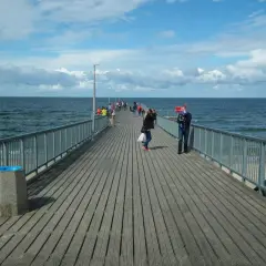 Ludzie spacerują po drewnianym deptaku nad morzem. Słońce przebija się przez chmury, a fale delikatnie uderzają o brzeg.