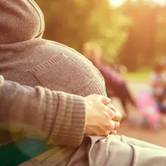 Gravid kvinna sitter på en parkbänk, njuter av solen. Hon håller sin mage, som om hon väntar på sitt barn. I bakgrunden syns andra människor och barnvagnar. Det är en varm och solig dag, med gula flytningar i luften.