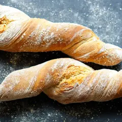Zwei gedrehte Brote, die an ein Wurzelbrot Rezept dunkel erinnern, liegen auf dunklem Untergrund, bestäubt mit Mehl.