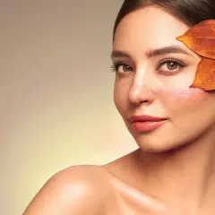 Une femme au teint légèrement rougi, cachant une partie de sa joue avec des feuilles d'automne. Une astuce pour savoir comment enlever des rougeurs rapidement.