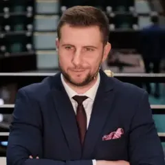 Mężczyzna w garniturze z krawatem i poszetką, siedzi na tle sali sejmowej.