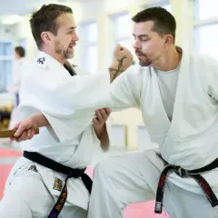 Två män i judodräkter tränar en judo kast. En av dem håller i ett träföremål.