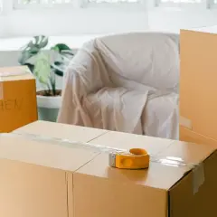 Cartons de déménagement prêts, dont un marqué "KITCHEN". Une valise et un fauteuil bâché complètent la scène.