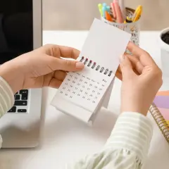 Ręce przeglądają kalendarz, szukając lipiec święta wolne od pracy. Obok laptop i kolorowy notatnik.