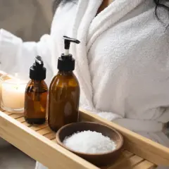 Kobieta w szlafroku trzyma tacę z kosmetykami: świecąca świeca, szczotka, butelki z płynami i sól do kąpieli. Idealny zestaw na spa dla niej.