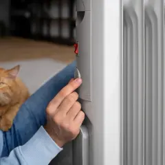 Persona ajusta un emisor térmico, comparándolo con un radiador de aceite, mientras acaricia a su gato.