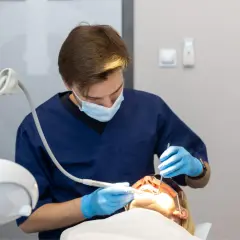 Dentysta w niebieskiej masce i rękawiczkach wykonuje zabieg. Pacjentka ma założone okulary ochronne. Nowoczesne znieczulenie komputerowe zapewnia komfort.