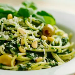 Pasta con pesto de pistacho, piñones, aceitunas verdes y queso rallado, decorada con hojas de albahaca fresca.