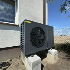 Nowoczesna pompa ciepła Geo Power, która obniża koszt ogrzewania domu. Urządzenie stoi na betonowych bloczkach przy ścianie budynku.