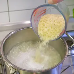 Reis im Topf kochen: Eine Hand schüttet Reis aus einem Messbecher in kochendes Wasser.