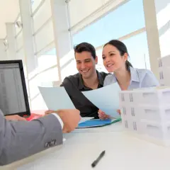 Ein Paar bespricht mit einem Berater Unterlagen, während Modellhäuser auf dem Tisch stehen. Der Berater absolviert vielleicht ein **Immobilienfachwirt berufsbegleitend** Studium.