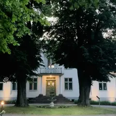Vitt hus med gröna dörrar och fönster, omgivet av träd. En fontän framför entrén. Drottning Victorias vilohem.