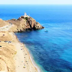 Costa de **Cabo de Gata** con faro en la cima de un acantilado, playa con gente y aguas cristalinas.