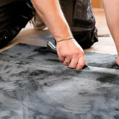 Mann schneidet mit einem Teppichmesser einen grauen Teppich zu. Das Verlegen des Teppichs wird präzise ausgeführt.