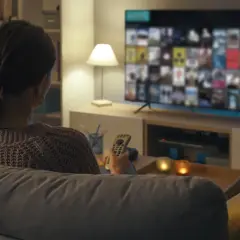 Kobieta z pilotem w ręku ogląda film na telewizorze, zastanawiając się, jak podłączyć komputer do telewizora, by cieszyć się większym ekranem.