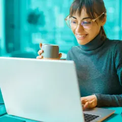 Uśmiechnięta kobieta z kubkiem kawy przegląda na laptopie informacje o dofinansowania na otwarcie firmy.
