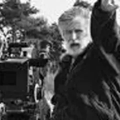 Edward Kłosiński, z siwą brodą, gestykuluje na planie filmowym. W tle widać kamerę i drzewa.