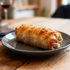 Une délicieuse crêpinette de porc dorée, enveloppée dans une pâte feuilletée croustillante, prête à être dégustée sur une assiette grise.
