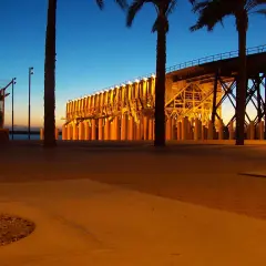 Atardecer en el parque de las almadrabillas, con palmeras y un puente iluminado sobre el mar.