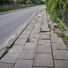 Zniszczony chodnik obok drogi. Płyty chodnikowe są popękane i nierówne, tworząc niebezpieczną ścieżkę dla pieszych.