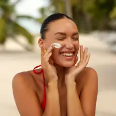 Uśmiechnięta kobieta na plaży nakłada krem z filtrem, który zawiera dwutlenek tytanu, chroniąc skórę przed słońcem.
